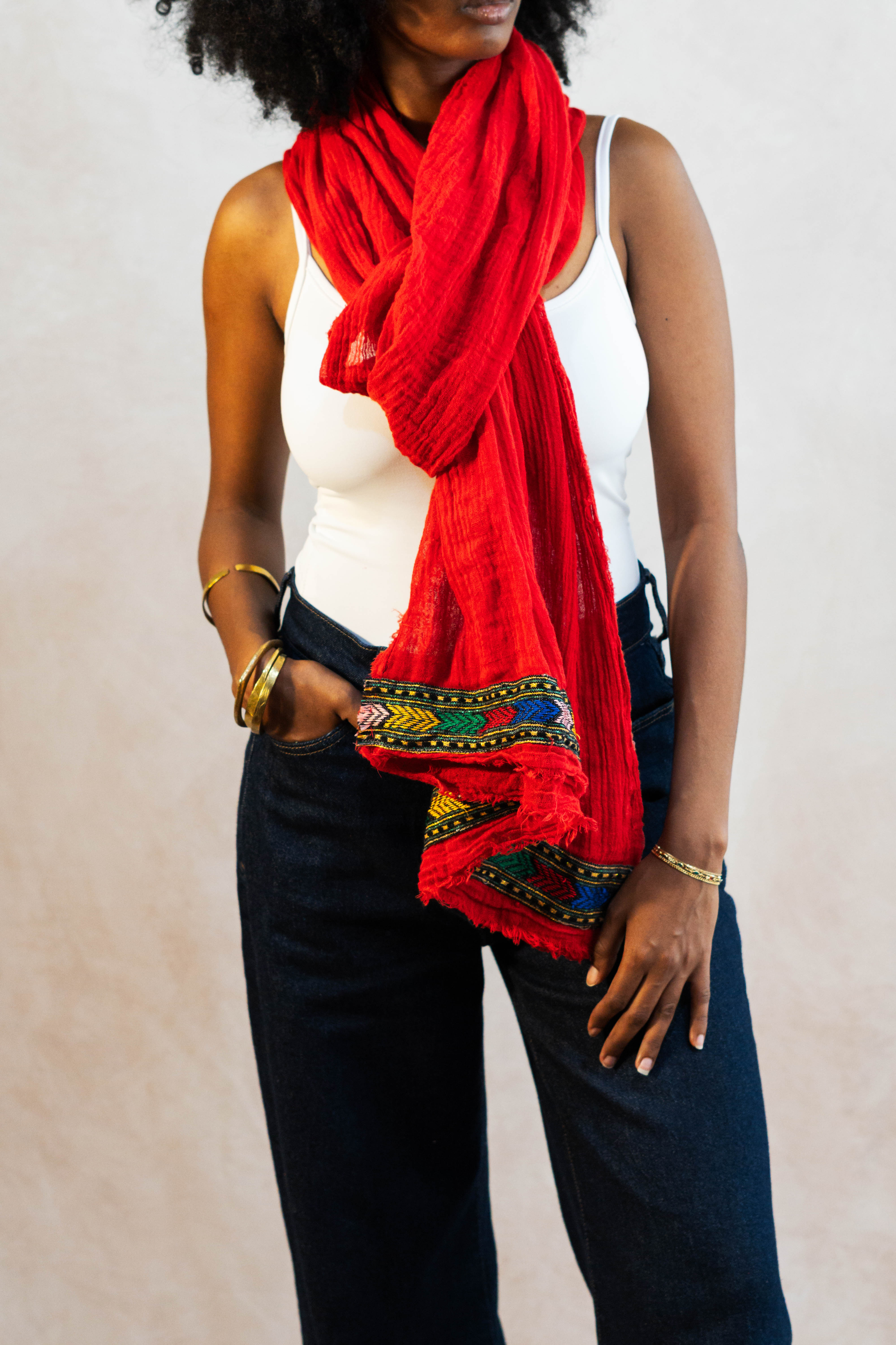A woman wearing a red hand-woven cotton scarf with intricate Tilet 
patterns, black pants, and gold jewelry against a plain background.