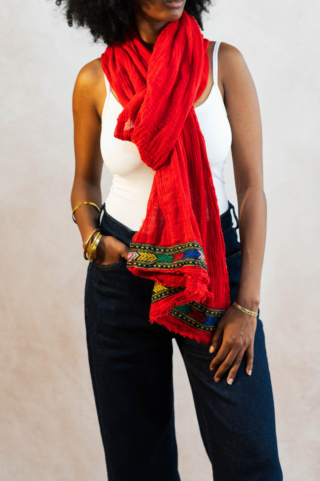 A woman wearing a red hand-woven cotton scarf with intricate Tilet 
patterns, black pants, and gold jewelry against a plain background.